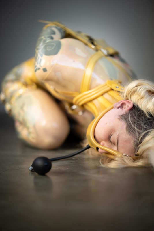 A sexy photograph of Cam Damage, in transparent latex. Tagged with: gagged, neck corset & armbinder. Posted April 2021.